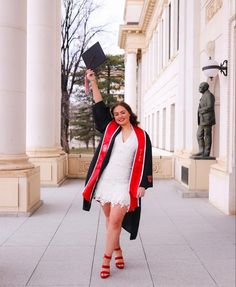 a woman in white dress and red jacket holding up a black graduation hat while standing on the sidewalk