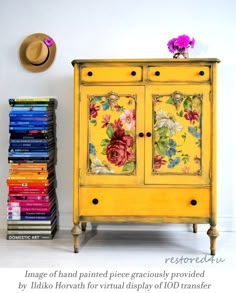 a yellow cabinet with flowers painted on the doors and drawers is next to a stack of books