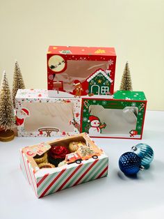 three boxes with christmas treats in them sitting on a table next to small trees and ornaments