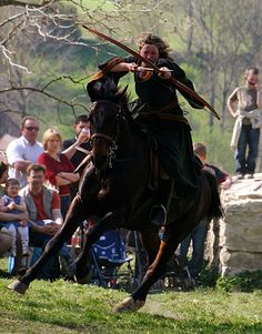 a woman riding on the back of a brown horse next to a forest filled with people