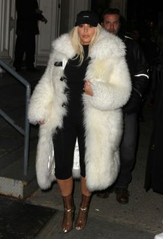 the blonde woman is wearing a white fur coat and black cap, while she's walking