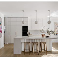 a white kitchen with three stools in front of an island and paintings on the wall