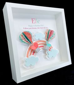 a white shadow box with some paper cut outs in the shape of a rainbow and clouds