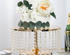 a wedding cake with white flowers and greenery on a gold stand next to champagne bottles