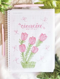 a notebook with pink flowers drawn on it next to green leaves and pencils in the background