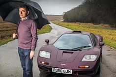 a man holding an umbrella standing next to a sports car on a road with grass and trees in the background