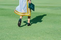 a woman in a dress is walking on the grass with a green hat and book