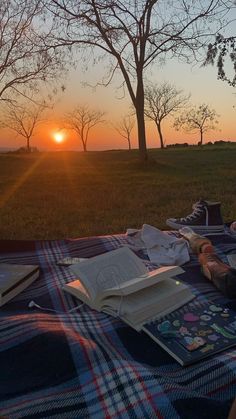 an open book sitting on top of a blanket next to a tree at sunset with the sun setting in the background