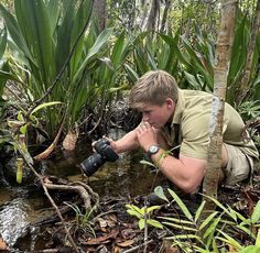 a man kneeling down in the water with a camera on his hand while looking at plants