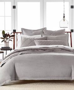 a bed with grey linens and pillows in a room next to two large windows