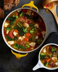 two bowls of soup with broccoli, carrots and other vegetables next to bread