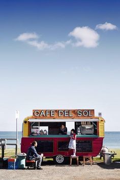 two people are standing at the front of a food truck by the ocean with their hands in their pockets