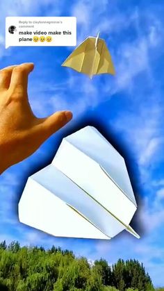 a hand is flying a paper airplane in the air with trees and blue sky behind it