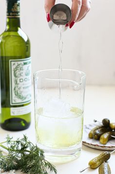 a person pouring water into a glass filled with ice and green olives next to bottles