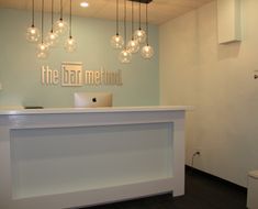 a white reception desk with lights hanging from it's ceiling and the words the bar method above it