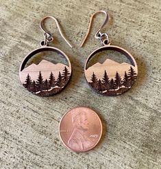 two earrings with trees and mountains on them, one has a penny in the foreground