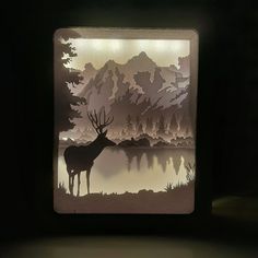 a deer standing in front of a lake surrounded by trees and mountains with lights on