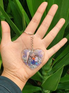 a hand holding a heart shaped keychain in front of green plants and leaves