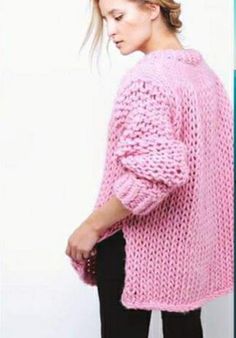 a woman standing in front of a white wall wearing a pink sweater and black pants