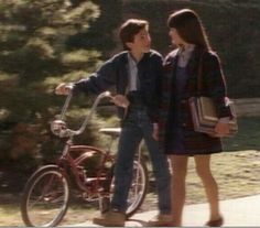 a young man and woman standing next to a bike on a sidewalk with trees in the background