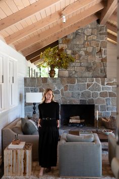 a woman standing in a living room next to a stone wall and fireplace with wood beams on the ceiling