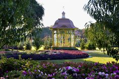 a gazebo surrounded by flowers and trees in the middle of a park with lots of greenery