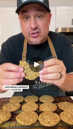 a man holding up some cookies on top of a cooling rack in front of a pile of muffins