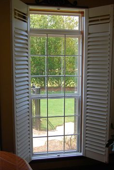 an open window with white shutters in front of a green yard and trees outside