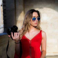 a woman wearing sunglasses holding a camera and looking at the camera with her hand in her pocket