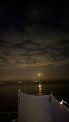 the moon is setting over the ocean with clouds in the sky