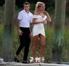 a woman in a white dress standing next to a man on a boat near tall grass