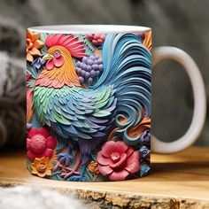 a colorful rooster coffee mug sitting on top of a wooden table next to a pile of yarn