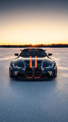 an orange and black sports car is on the ice in front of some trees at sunset