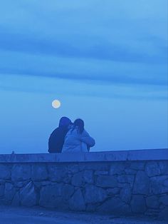 two people sitting on a stone wall looking at the moon