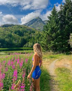 a woman standing in the middle of a field with purple flowers and mountains behind her