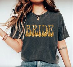 Shine in style with this elegant Western-style 'Bride' shirt featuring a glittery gold print. Perfect for your bridal party and making your special day even more memorable as you say 'I do.' Our garments are pre-shrunk and soft-washed for perfect fit and supreme comfort. The fabric is garment-dyed, imbuing each shirt with a deep, eye-catching hue. Set-in sleeves offer a traditional and enduring style. Enhanced durability comes from the double-needle stitching on the sleeves and bottom hem. Twill Bachelorette T Shirts, Special Occasion Jewelry, Bride Shirt, Halo Hair, Floral Bracelet, Back Necklace, Bride Shirts, Glitter Gold, Bohemian Necklace