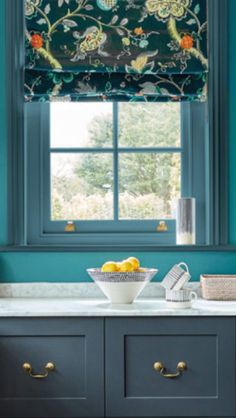 a bowl of fruit is on the counter in front of a window with blue walls