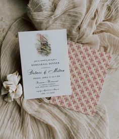 the wedding card is laying on top of some fabric next to a white and pink flower