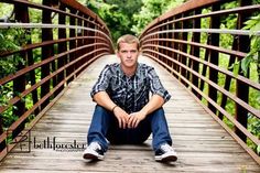 a young man is sitting on a bridge in the woods and posing for a photo