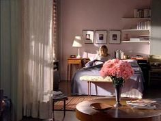 a woman sitting on a bed in a bedroom next to a table with pink flowers