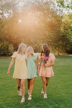 three girls walking in the grass with their arms around each other and one girl holding flowers