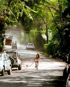 a woman is walking her dog down the street while cars are parked on the side of the road