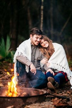 a man and woman sitting in front of a campfire smiling at the camera with their arms around each other