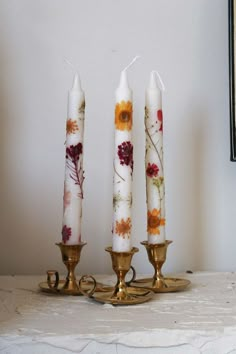 three white candles with flowers on them sitting on a marble shelf next to two brass candle holders