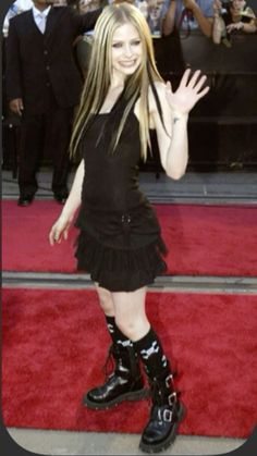 a woman in black dress and boots waving at the camera on red carpet with people behind her