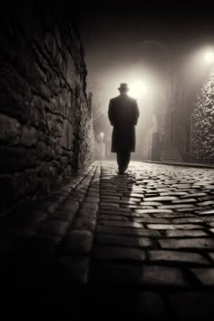 a man in a long coat and hat walks down a cobblestone street at night