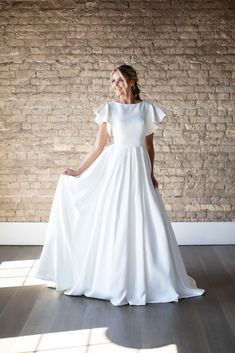 a woman in a white wedding dress standing against a brick wall with her hands on her hips