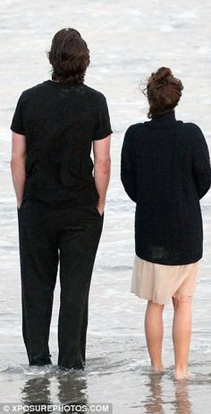 two people standing in the water looking out at the ocean and one person is wearing black