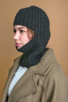 a woman wearing a gray hat and coat with a scarf around her neck, standing in front of a brown wall