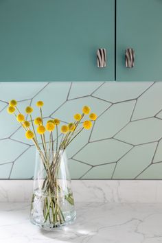 some yellow flowers in a clear vase on a marble counter top with blue cabinets behind it